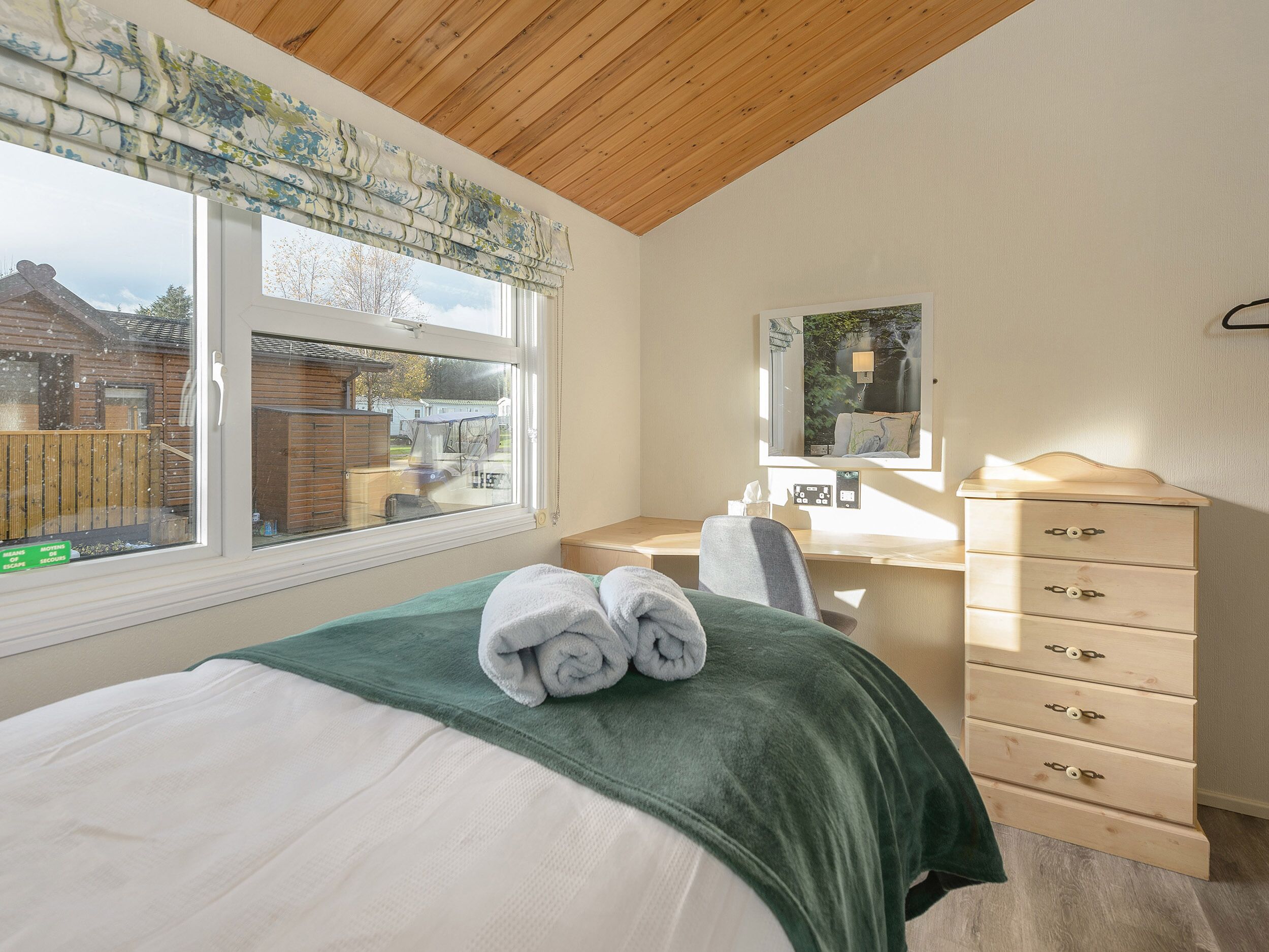 A bedroom with a bed, a desk, a dresser, a window with a view of the outside, and a mirror on the wall.