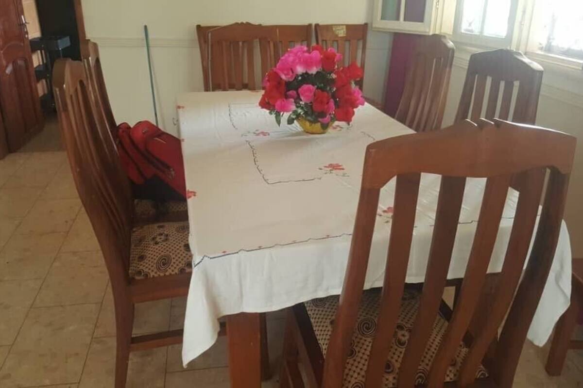 A dining area with a table set for four, surrounded by wooden chairs. A vase with flowers is placed on the table.