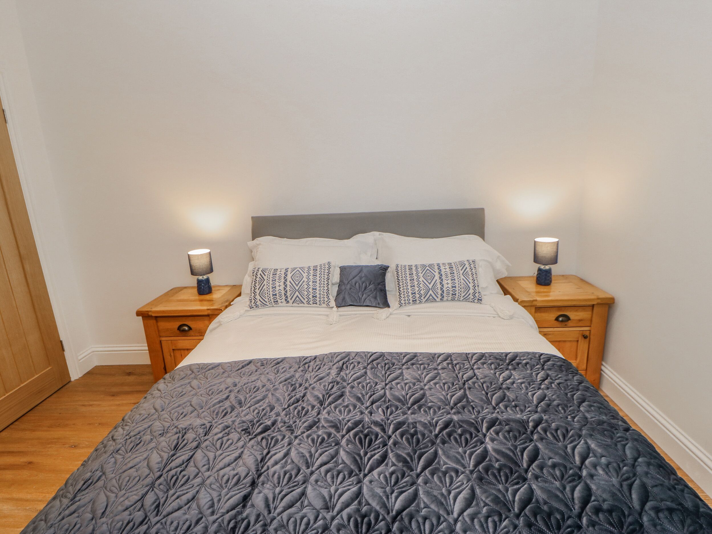 Una cama bien hecha con un edredón estampado, dos mesitas de noche con lámparas y suelo de madera.