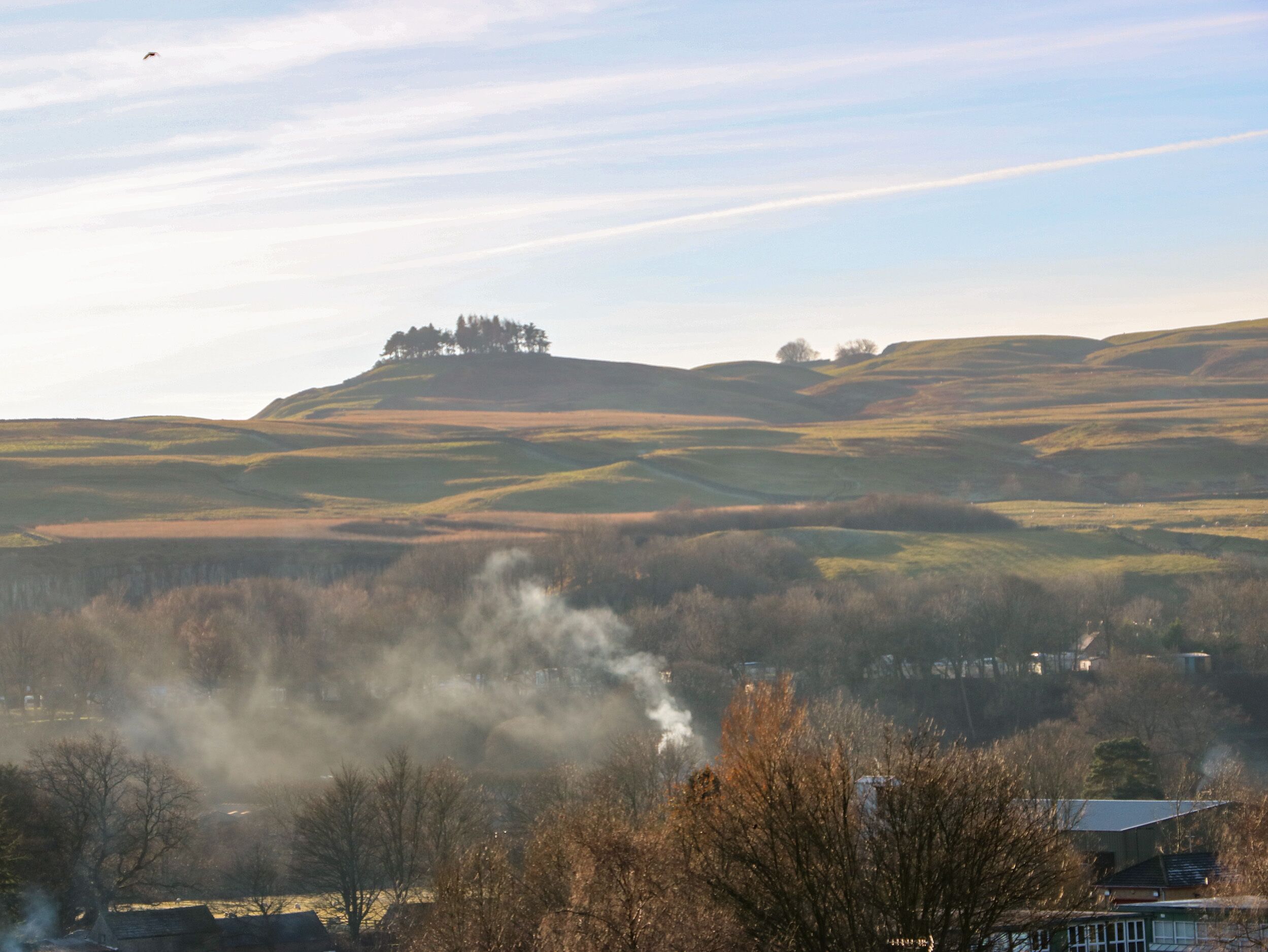 A landscape with rolling hills, a hilltop with trees, and a building emitting smoke.