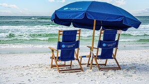 Dos sillas de playa bajo una sombrilla azul con el texto "LA DOLCE VITA" en ellas, ubicadas en una playa arenosa con el océano al fondo.