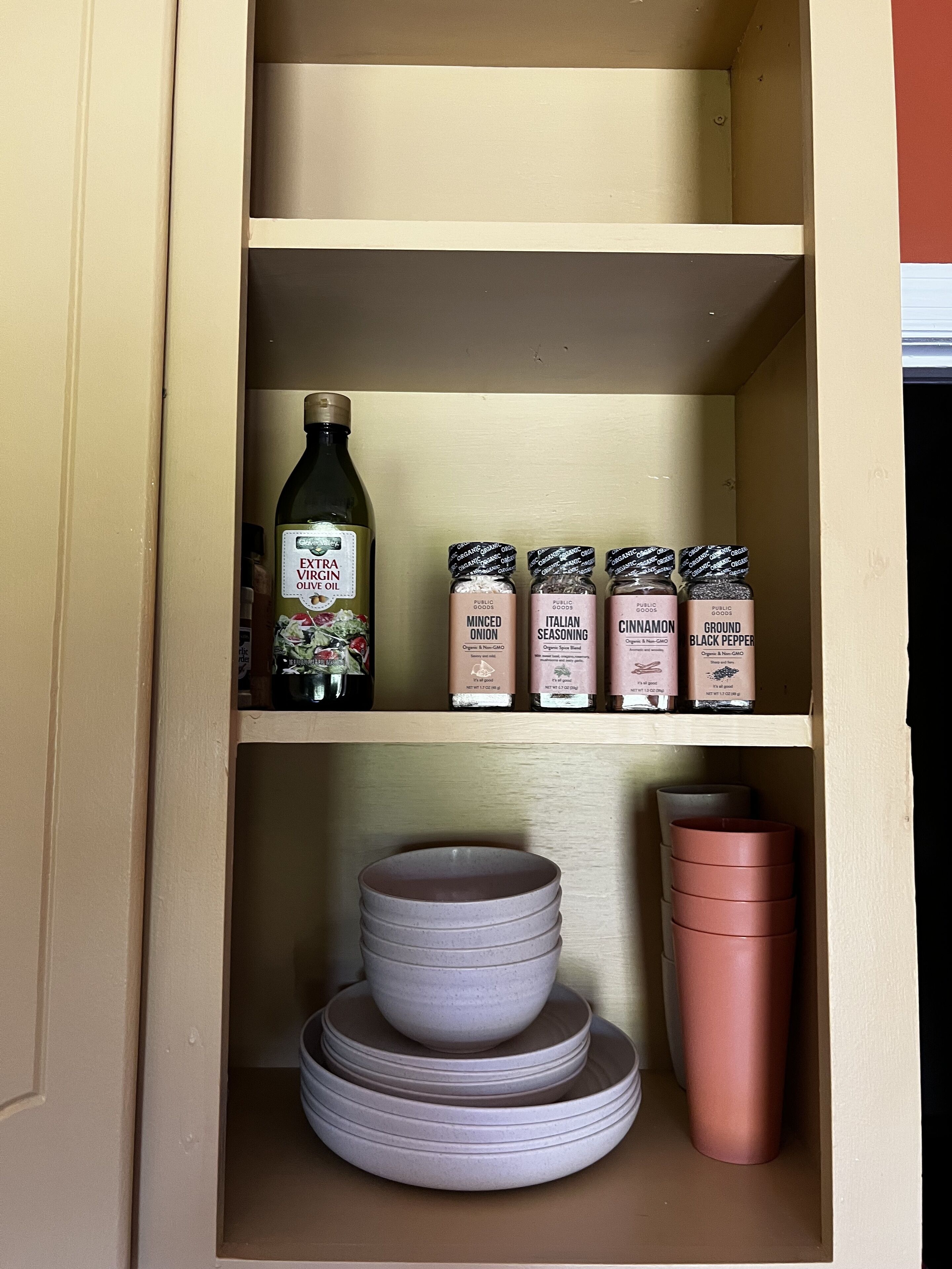 Un mueble de cocina con estantes que contiene una botella de aceite de oliva virgen extra, tarros de cebolla picada, hierbas italianas, canela y pimienta negra. Debajo de los estantes, hay platos apilados y una taza rosa.
