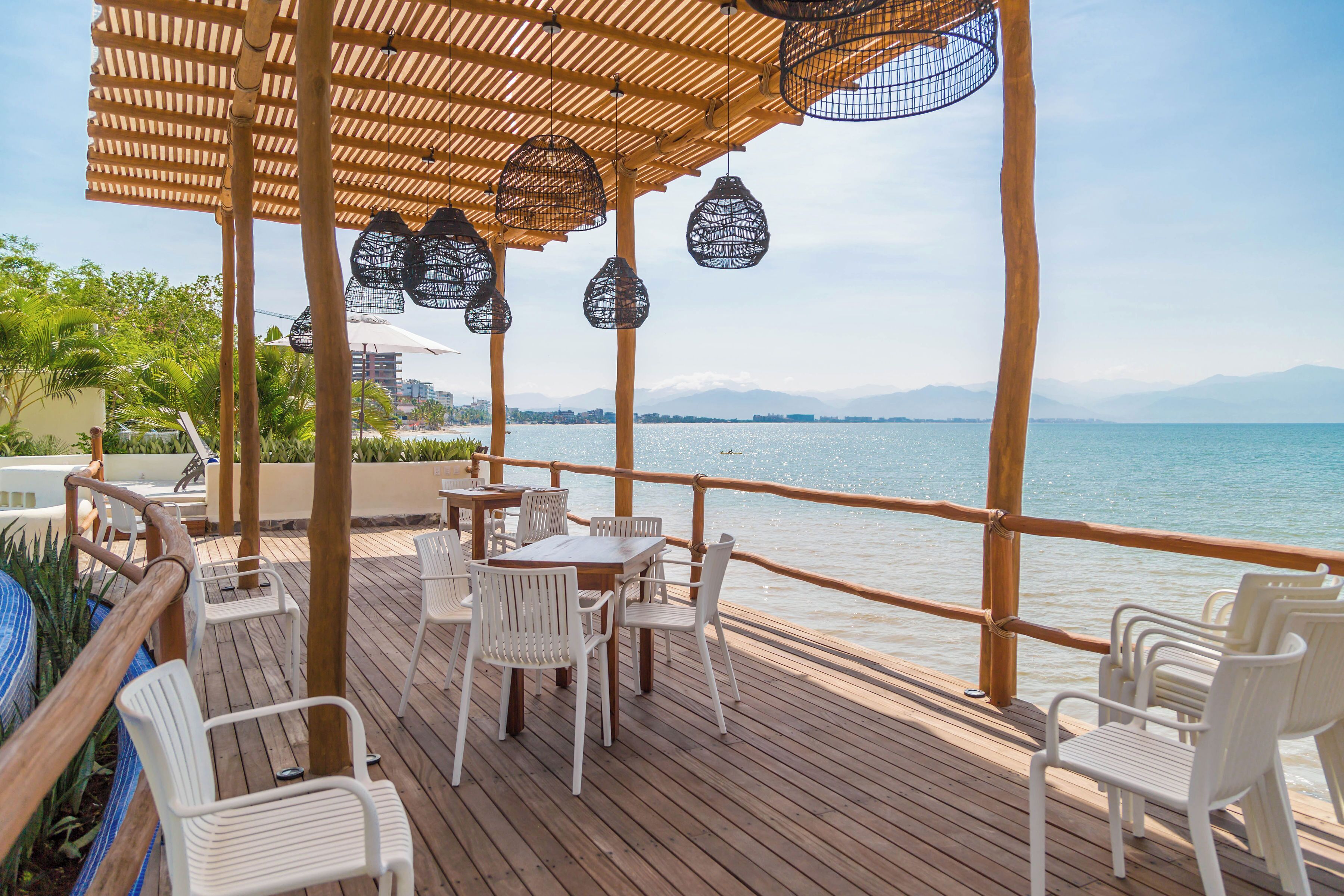 A wooden deck with white chairs and tables, overlooking a body of water with mountains in the distance.
