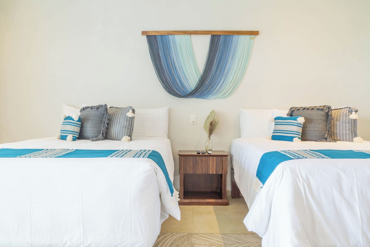 Une chambre d’hôtel avec deux lits, chacun agrémenté de draps blancs et de touches de bleu, une table de chevet en bois et une décoration murale tissée.