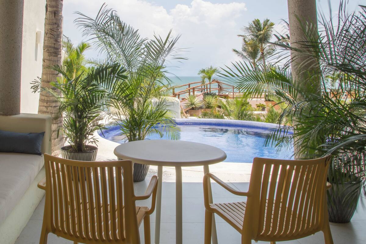 Un espace aménagé au bord de la piscine, avec une table et des chaises, entouré de plantes en pot et de palmiers, offrant une vue sur une piscine aux eaux d’un bleu limpide et un pont en bois.