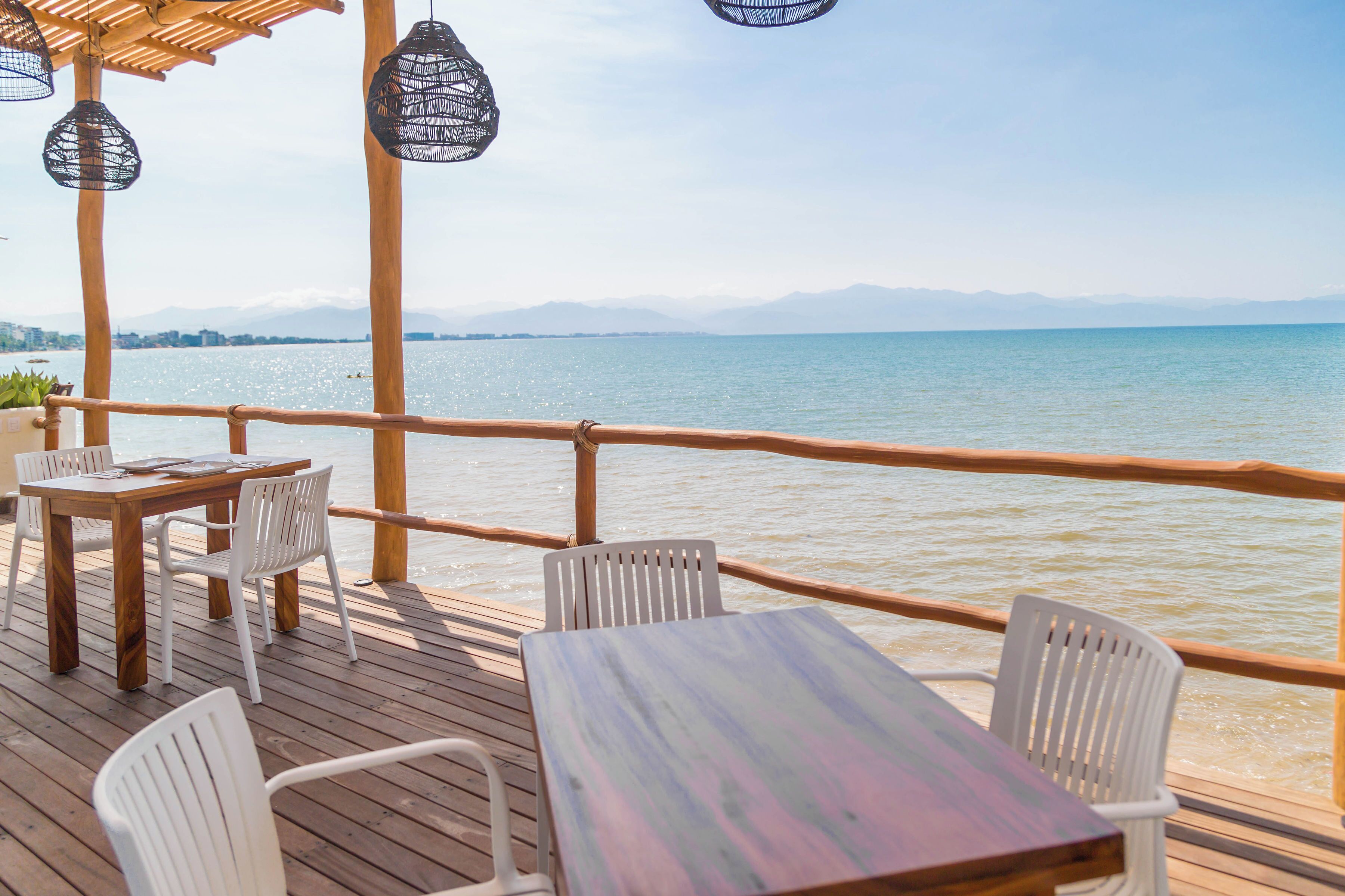 Une terrasse en bois, agrémentée de tables et de chaises, qui offre une vue sur une étendue d’eau, avec des montagnes à l’horizon.