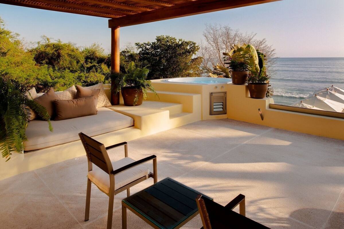 Un espace extérieur ensoleillé, aménagé avec des sièges, offrant une vue sur la mer, ainsi qu’un jacuzzi et des plantes en pot.