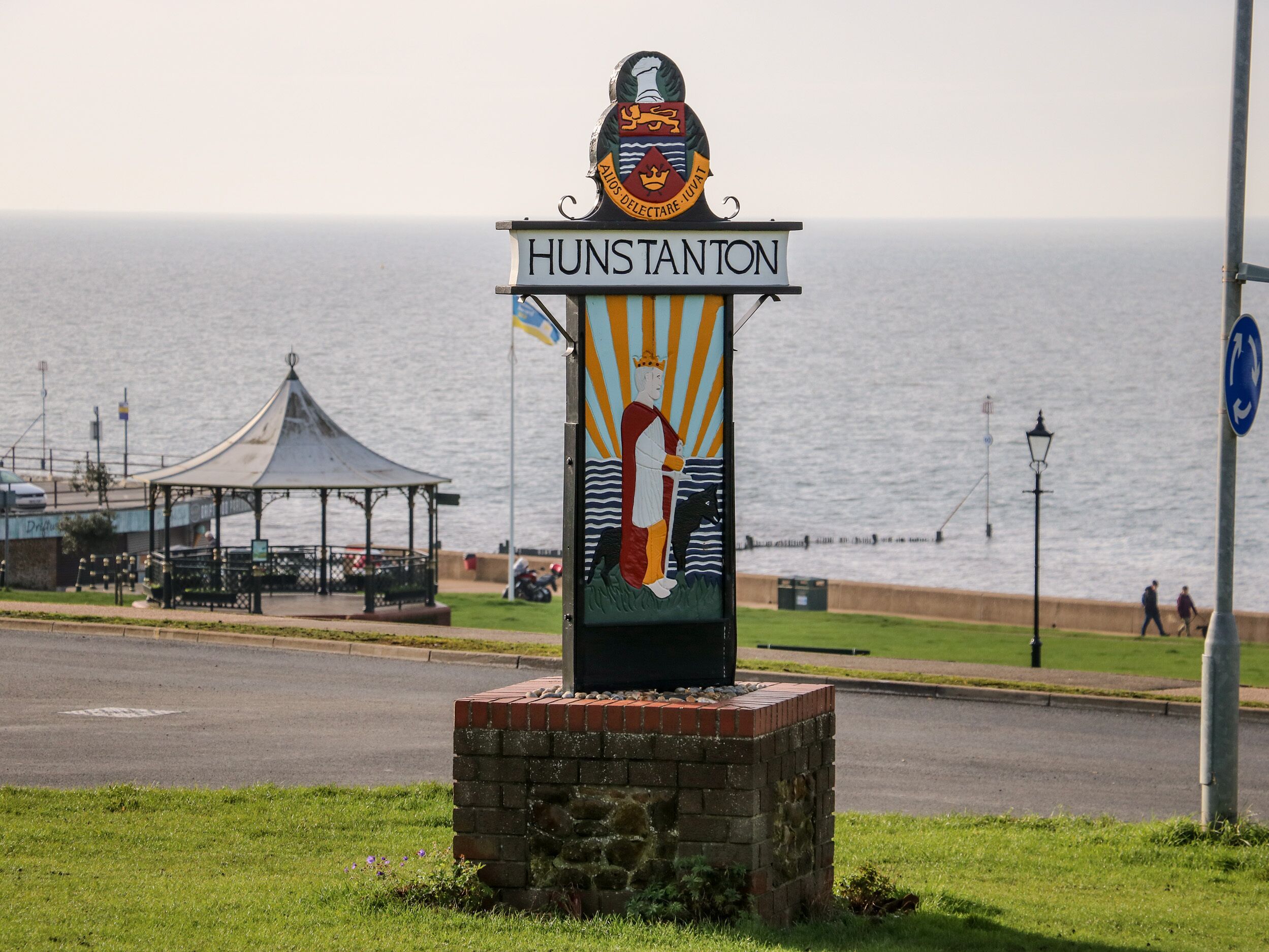 A sign with 'HUNSTANTON' and a coat of arms, located near a waterfront with a gazebo and a road.