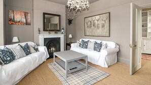 A living room with a fireplace, two white sofas with patterned cushions, a chandelier, and artwork on the walls. - Troon Cottage (Hunstanton)