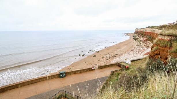 A coastal walkway with a concrete barrier, a sandy beach, and a cliff with vegetation. - Troon Cottage (Hunstanton)