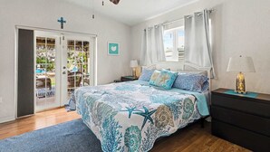 Una camera da letto con un letto, una ventola a soffitto, un comodino, una lampada, una finestra con tende e una porta che dà su un balcone, oltre a una croce appesa al muro.