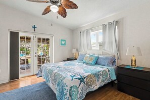 A bedroom with a bed, a ceiling fan, a nightstand, a lamp, a window with curtains, a door leading to a balcony, and a cross on the wall.