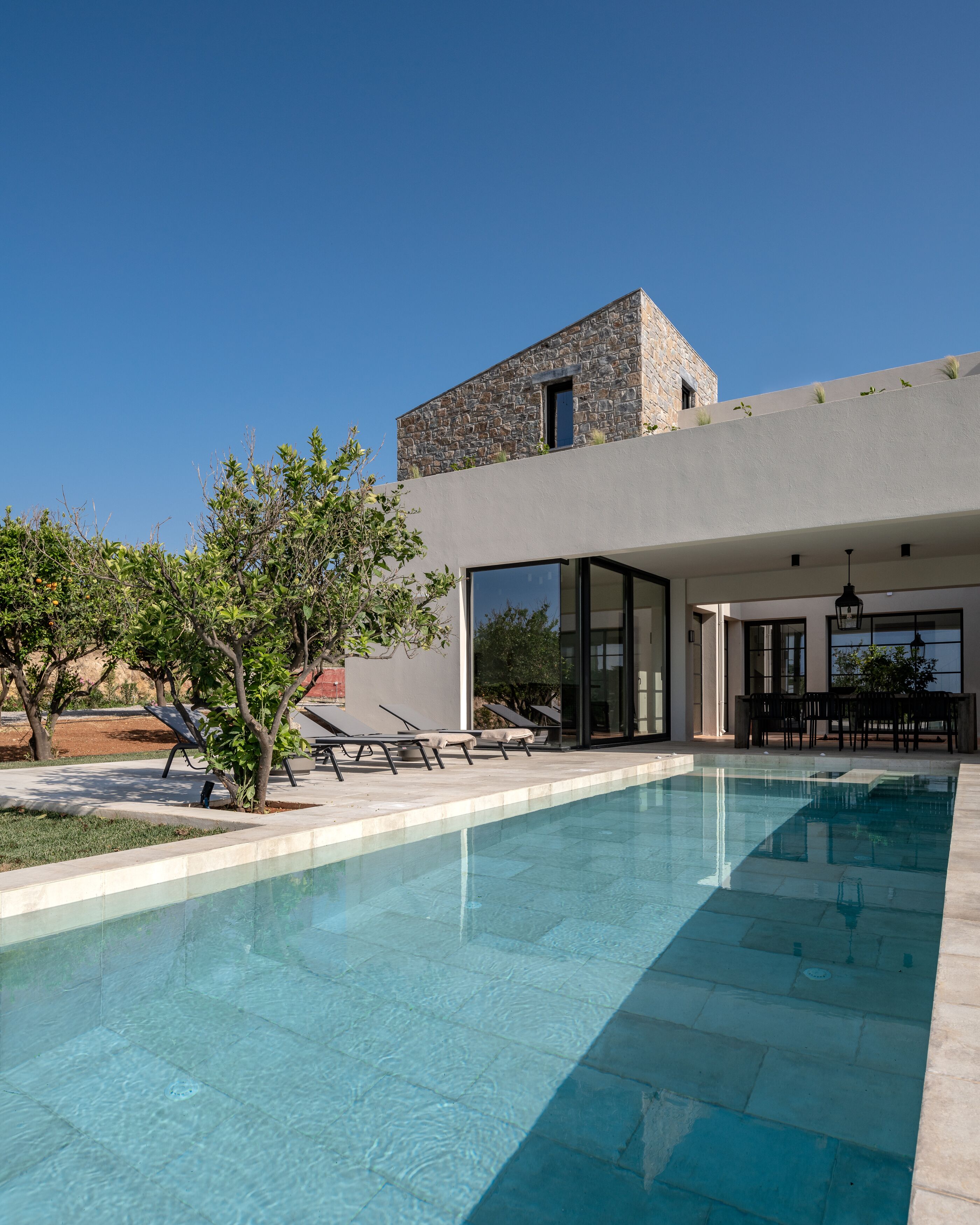 A modern pool area with clear blue water, lounge chairs, and a stone building with a flat roof.