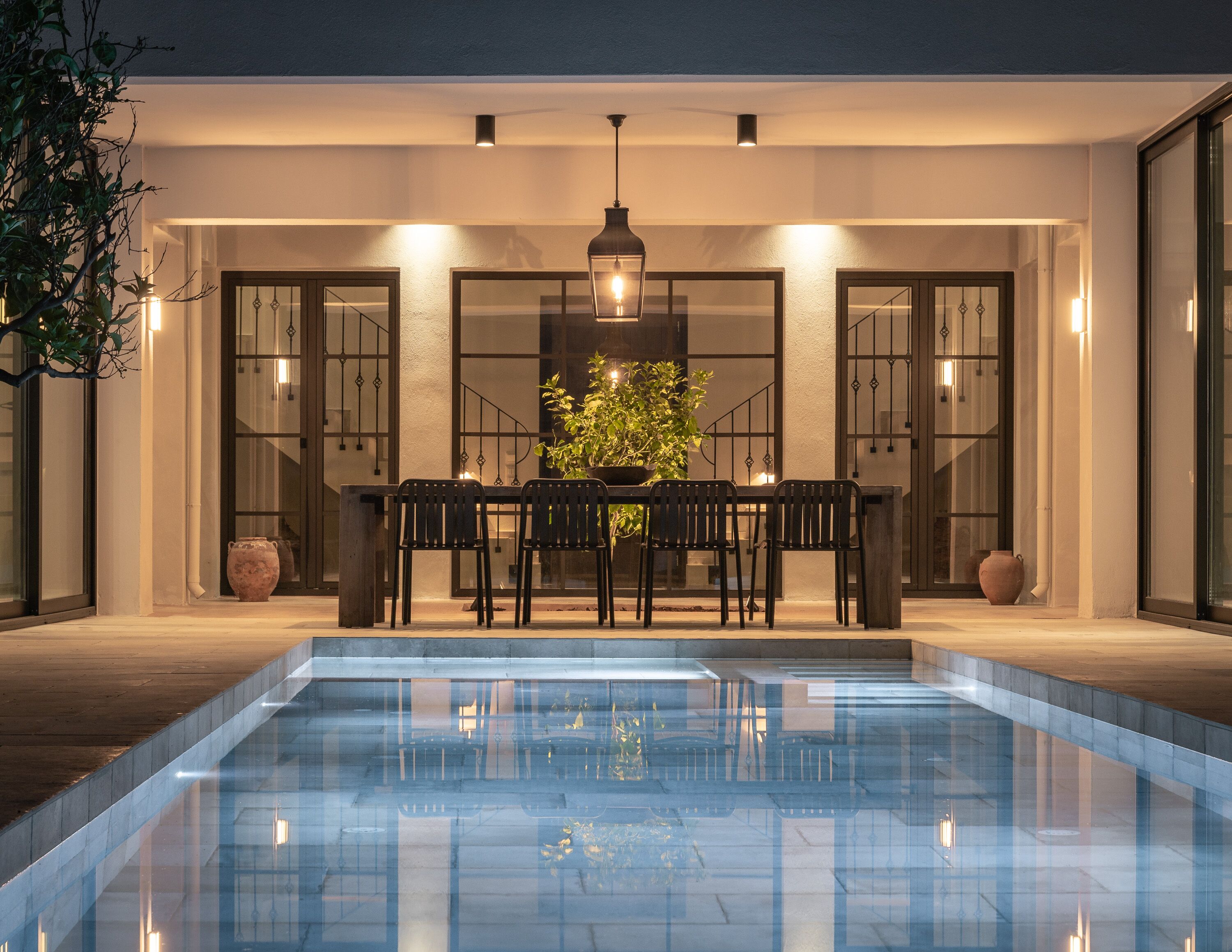 An outdoor pool area with a dining table and chairs, surrounded by walls with large windows and doors.
