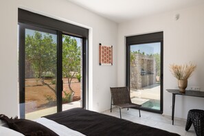 A modern bedroom with a large sliding glass door leading to an outdoor area, a wicker chair, and a small table with a vase. - Citrus Grove (Rethymno)