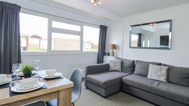 A modern living room with a grey sofa, a dining table set with plates and glasses, and a large window with curtains. - California Summers (Great Yarmouth)