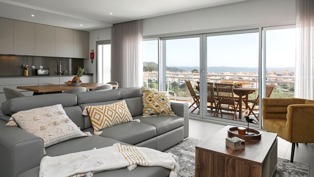 A modern living room with a grey sofa, wooden coffee table, and a city view through large windows. - Casa Urlaub - Panoramic Rooftop View (Nazaré)