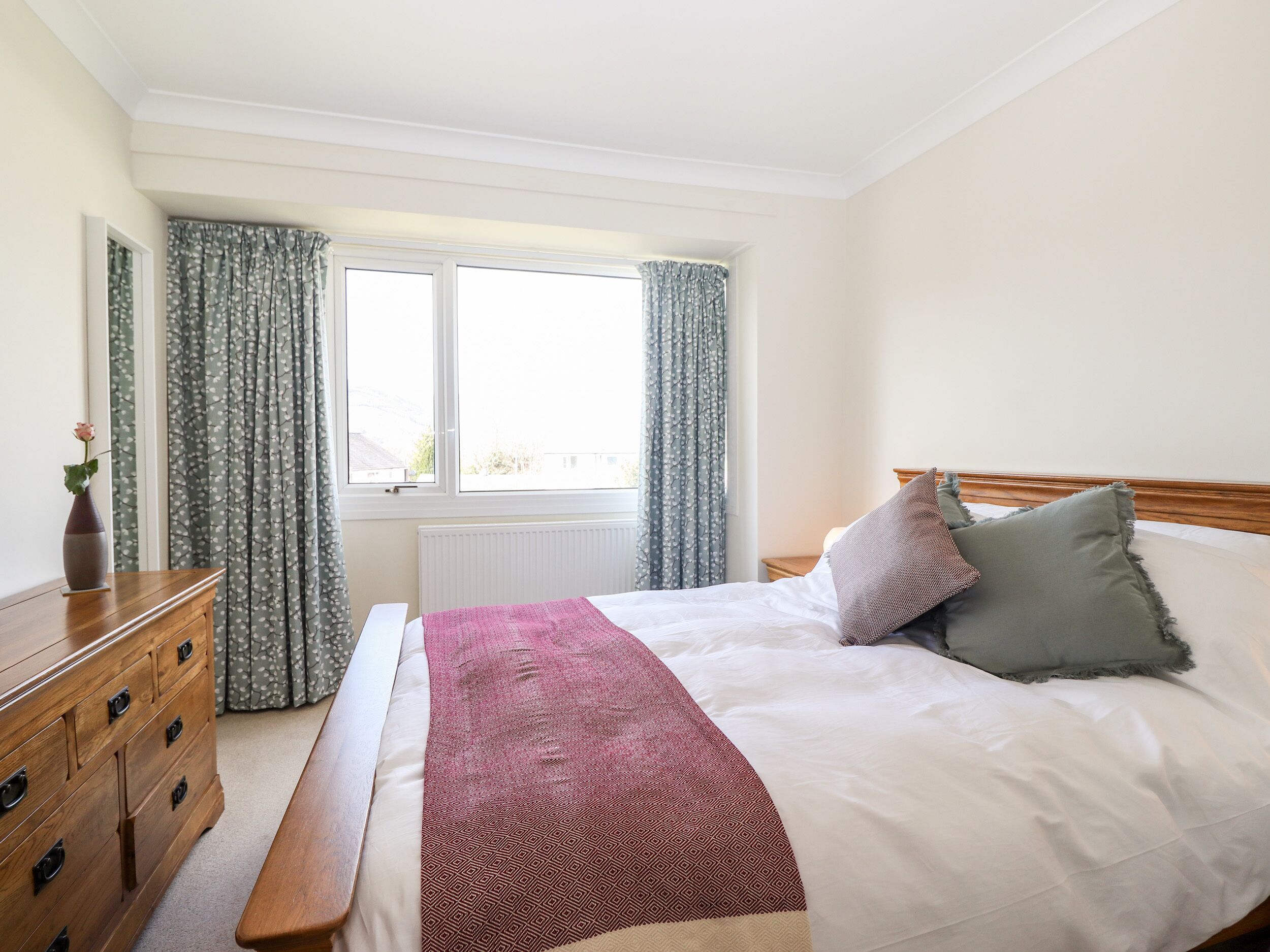 A bedroom with a bed, a wooden dresser, a window with curtains, and a radiator.