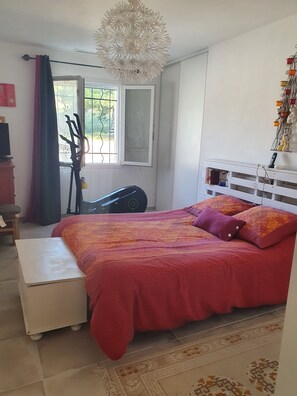 A bedroom with a large bed, a white bedside table, a window with bars, a ceiling light fixture, and a wall-mounted shelf with items.