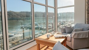 A room with large windows overlooking a marina with boats. There is a wooden table with a cup and a magazine. - 33 Dart Marina (Dartmouth)