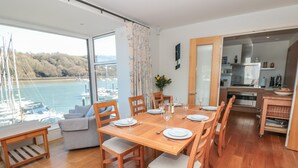A dining area with a wooden table set for six, large windows overlooking a marina, and a modern kitchen in the background. - 33 Dart Marina (Dartmouth)