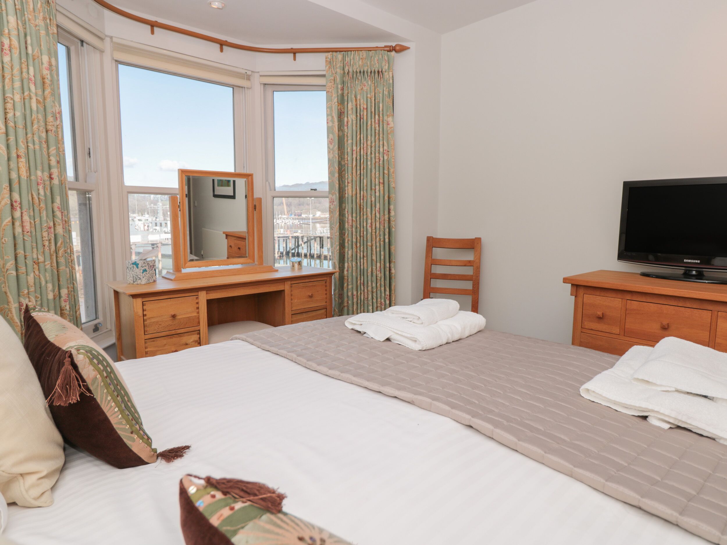 A hotel room with a large bed, a wooden desk, a chair, a television, and a window with curtains.