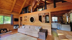 A spacious cabin with a high ceiling, a cozy living area with a sofa and a fireplace, a kitchen area, and a loft space with a window overlooking the outdoors. - Romantic Off-Grid Cabin (Lytton)