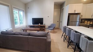 Ein modernes Wohnzimmer mit Sofa, Fernseher, Essbereich mit Barhocker und einer Küche inklusive Kühlschrank.