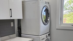 Ein Waschraum mit übereinander gestapelten Waschmaschinen, einem Waschbecken, Schränken und einem Fenster mit Blick auf Grünflächen.