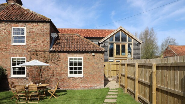 A brick house with a tiled roof, a wooden fence, and a grassy yard with outdoor furniture. - Acorn Cottage (Driffield)