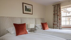A hotel room with two beds, a headboard, a nightstand, a lamp, a framed picture on the wall, and a window with curtains. - Acorn Cottage (Driffield)