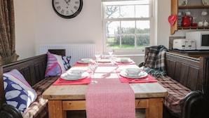 A dining area with a wooden table set for four, a clock on the wall, and a view of the outdoors through a window. - Acorn Cottage (Driffield)