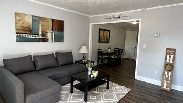 A living room with a sectional sofa, a coffee table, and a rug. There is a painting on the wall and a welcome sign.