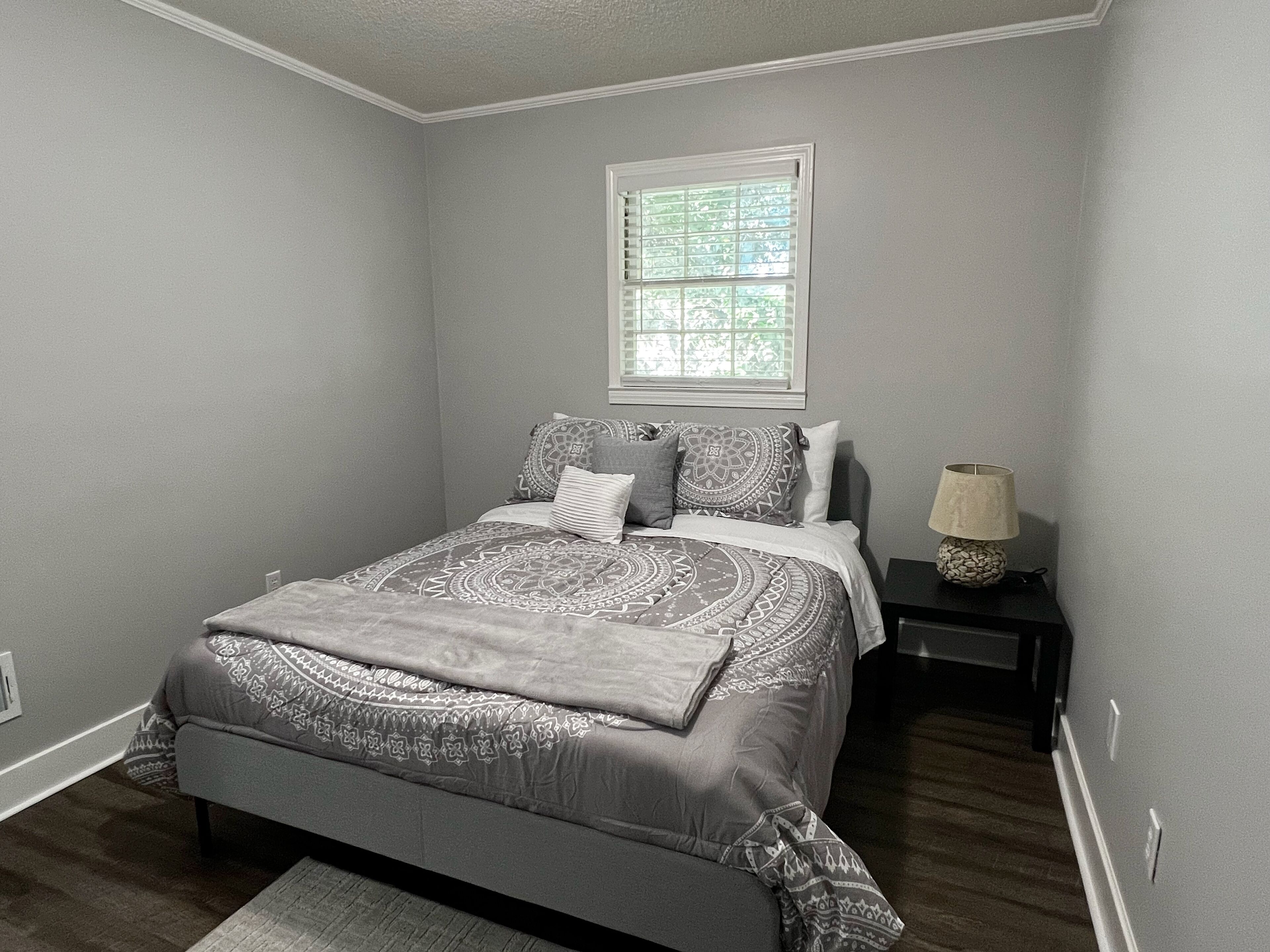 Un dormitorio con cama, mesita de noche, lámpara y ventana.