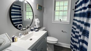 Une salle de bain avec un miroir rond, un lavabo avec des articles de toilette, des toilettes et une fenêtre donnant sur un espace verdoyant.