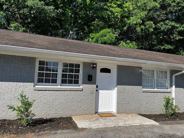 A single-story house with a grey brick exterior, a white front door, and a small front yard with two young trees. - Cozy Apt. Near Downtown (2BR, 1BA, 3 beds) (Greenville)