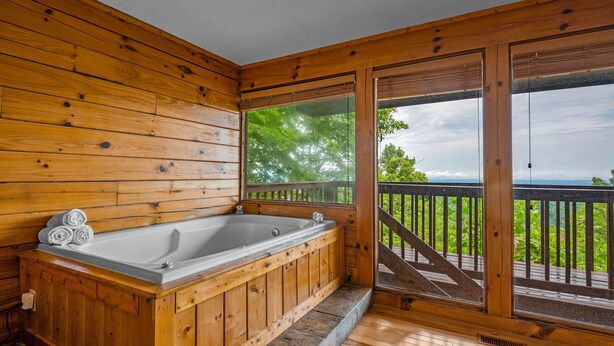 Eine Holzhütte mit einer großen Holzwanne, einem Balkon mit Geländer und Blick auf Bäume und Himmel.