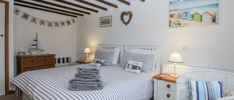 A bedroom with a bed, two bedside tables, a nightstand, a wicker chair, and a wall with a picture and a heart decoration.