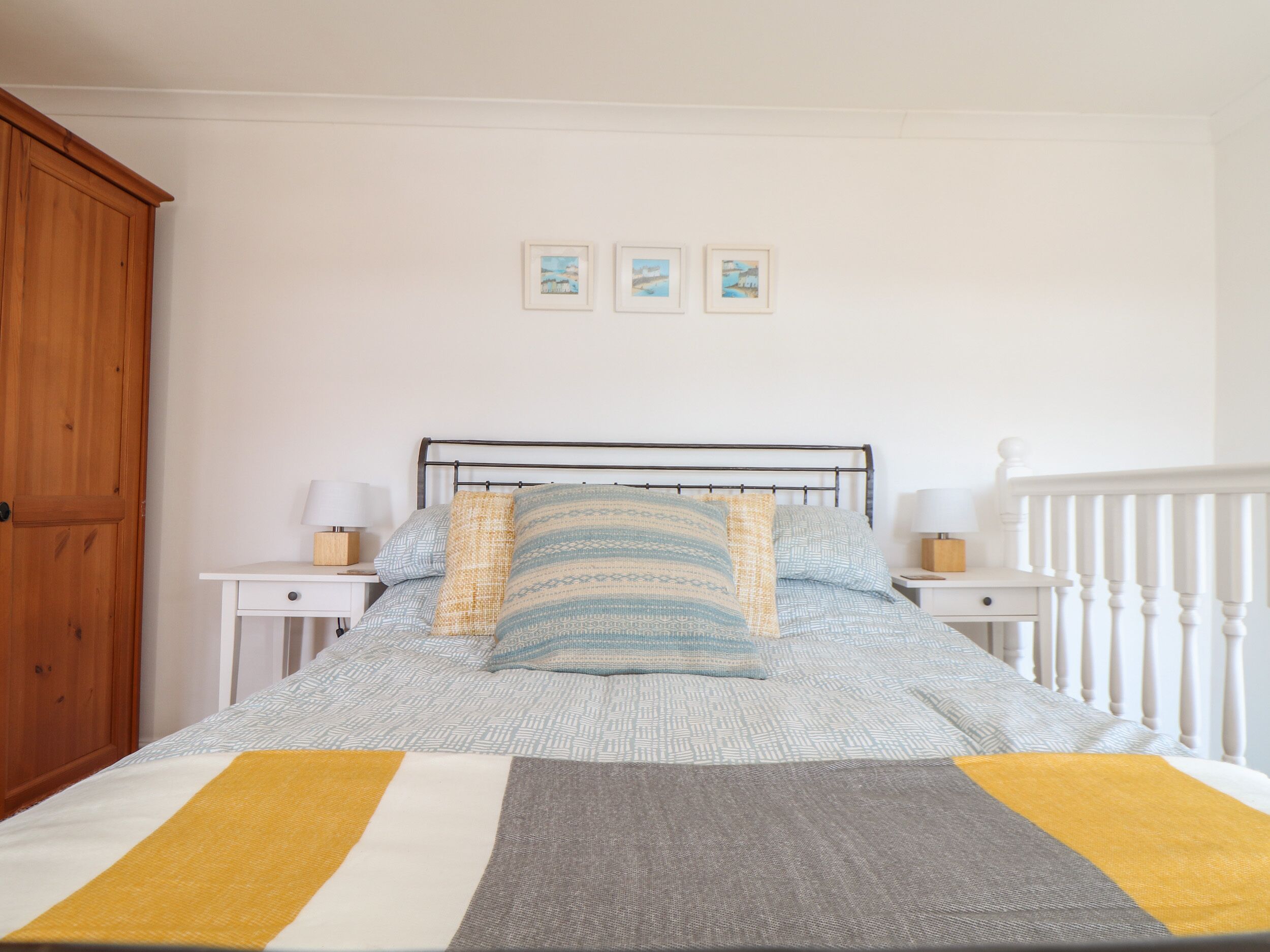 A neatly made bed with patterned bedding, two bedside tables with lamps, a wooden wardrobe, and three framed pictures on the wall.