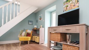 Ein Wohnzimmer mit Holzboden, einem gelben Sofa, einem runden Holztisch, einem Fernseher auf einem Holzschrank, Körbwäsche und einer Treppe.