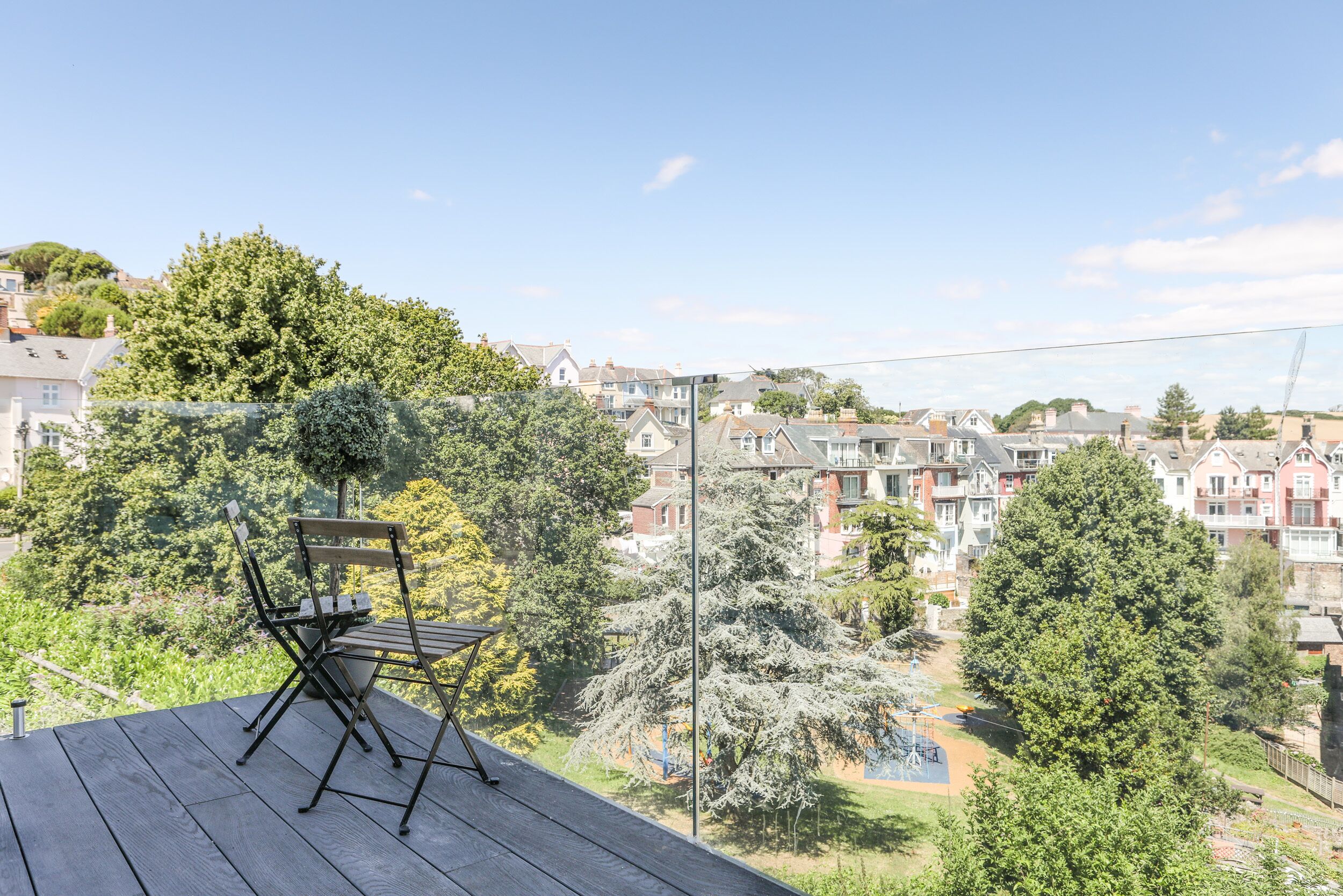 Un balcón con suelo de madera y una silla de metal, con vistas a una zona residencial con casas y árboles.