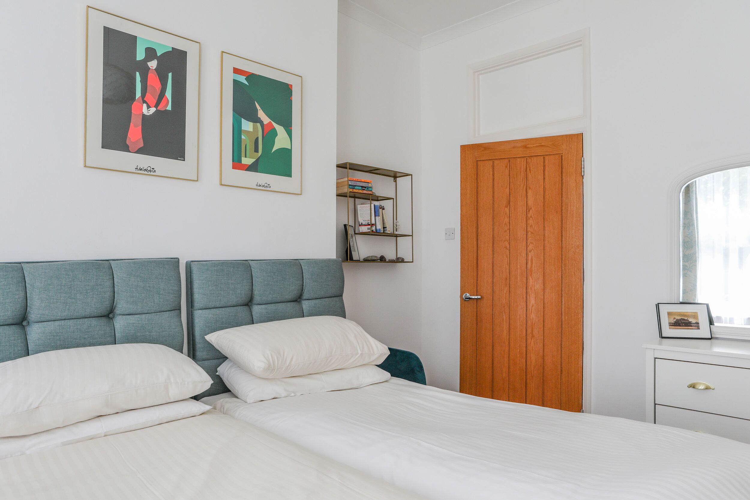 A bedroom with a bed, two framed artworks, a wooden door, and a window.