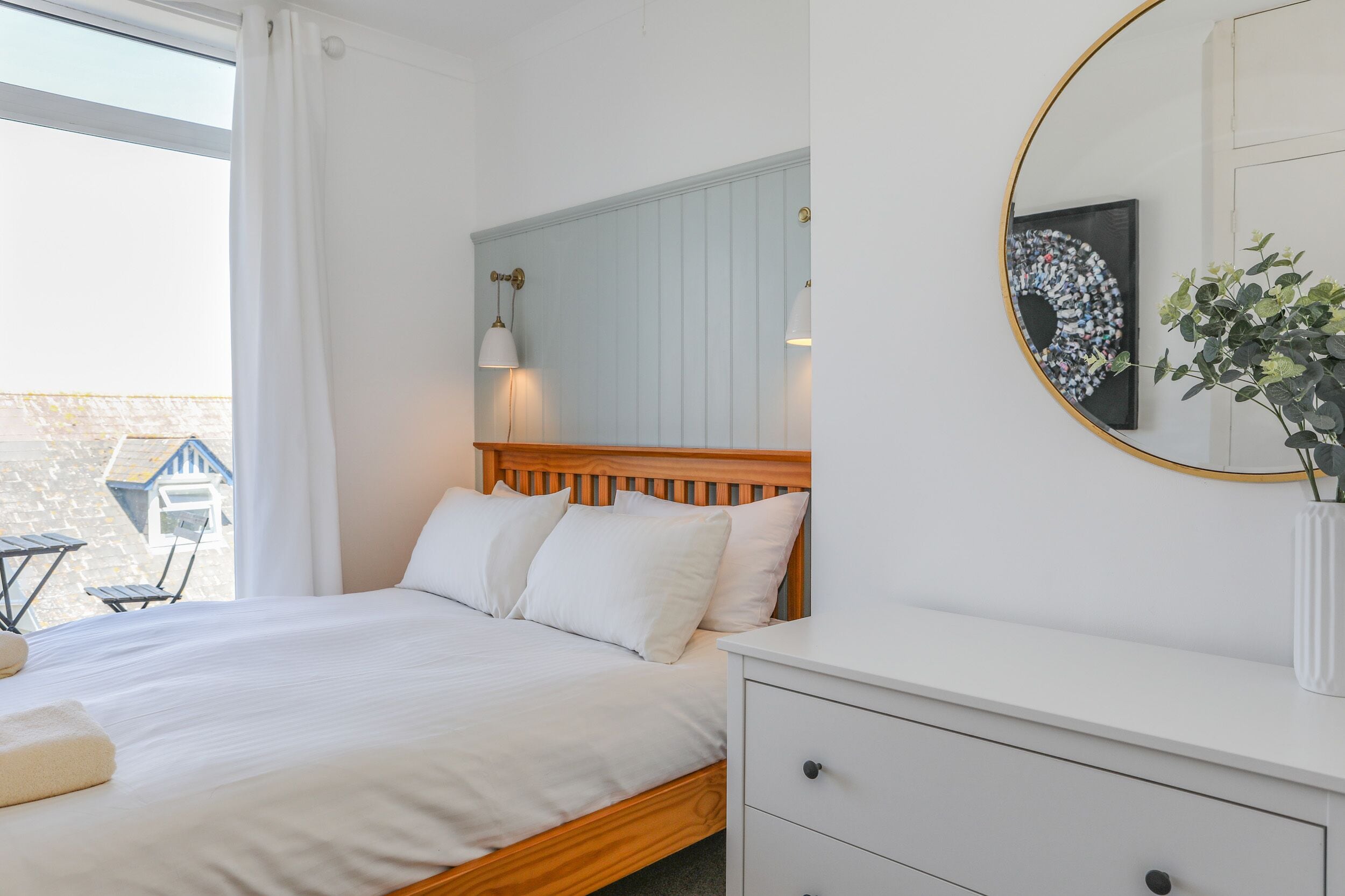 Une chambre à coucher avec un lit en bois, du linge de lit blanc, une table de chevet, un miroir et une fenêtre avec des rideaux.