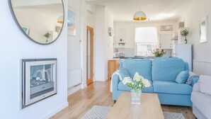 Una sala de estar moderna con un sofá azul, una mesa de centro de madera con flores, una chimenea y una cocina al fondo.