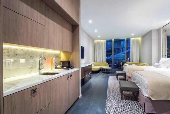A modern hotel room with a kitchen area, a sofa, two beds, and a view of the city at night.