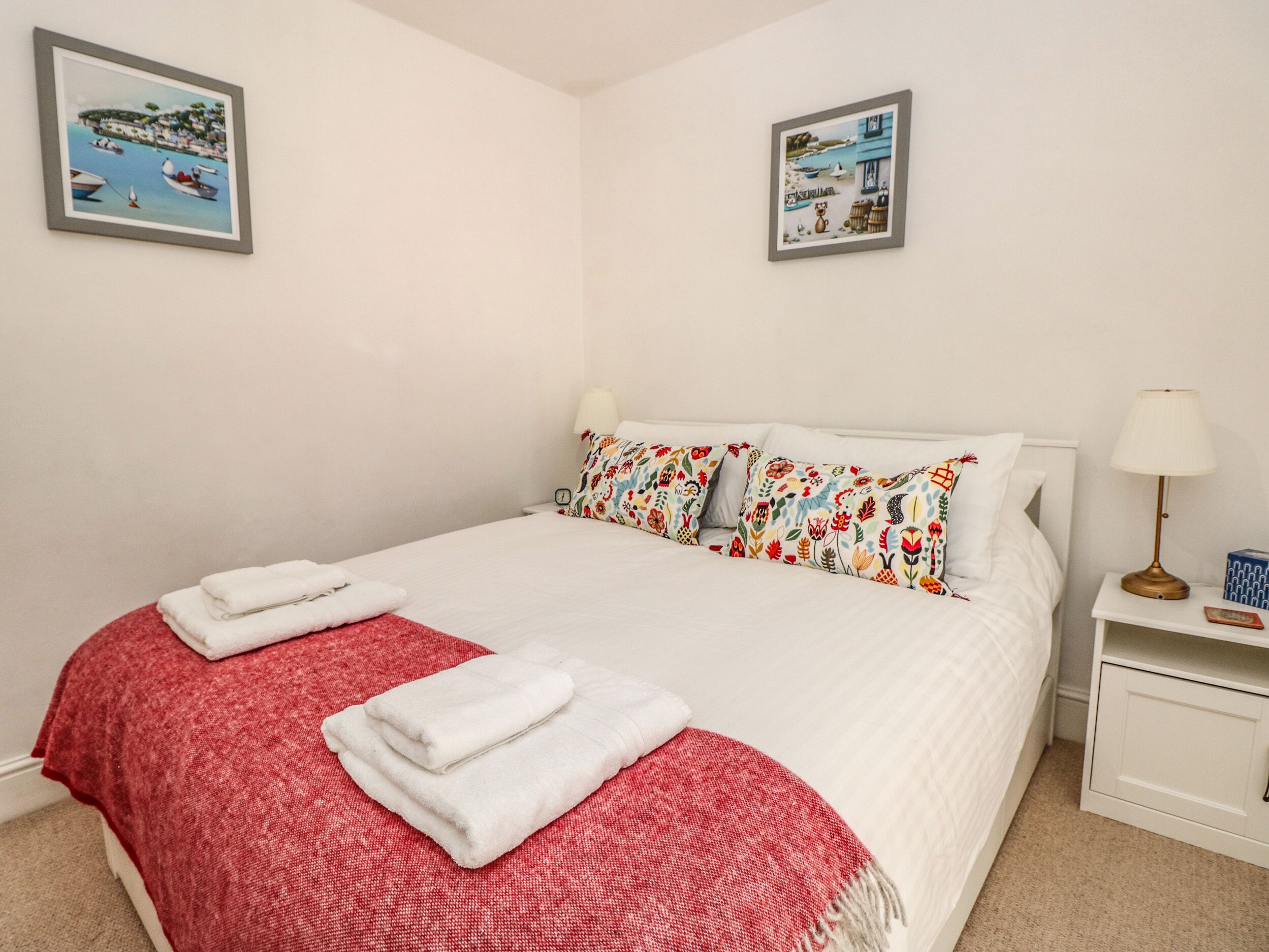 A neatly made bed with white linens, a red and white patterned throw, and two white towels. There are two wall-mounted pictures depicting coastal scenes.