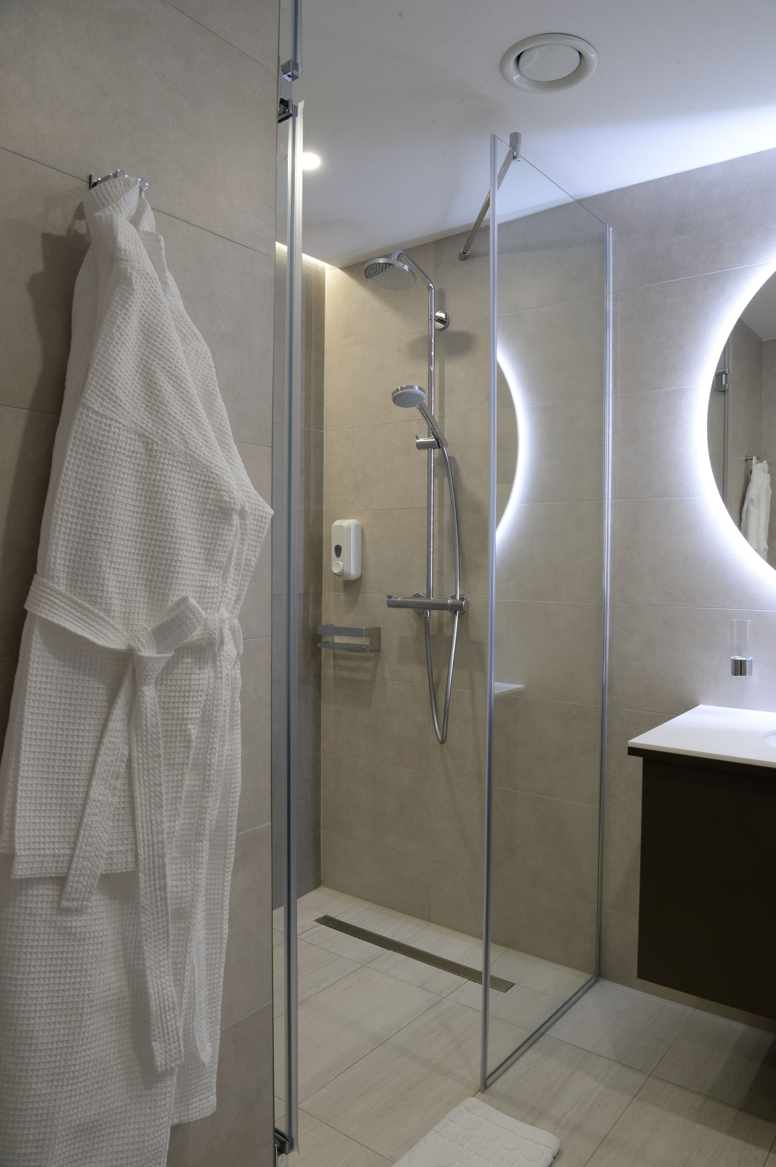 A modern bathroom with a glass-enclosed shower, a white bathrobe hanging on the wall, and a round mirror.