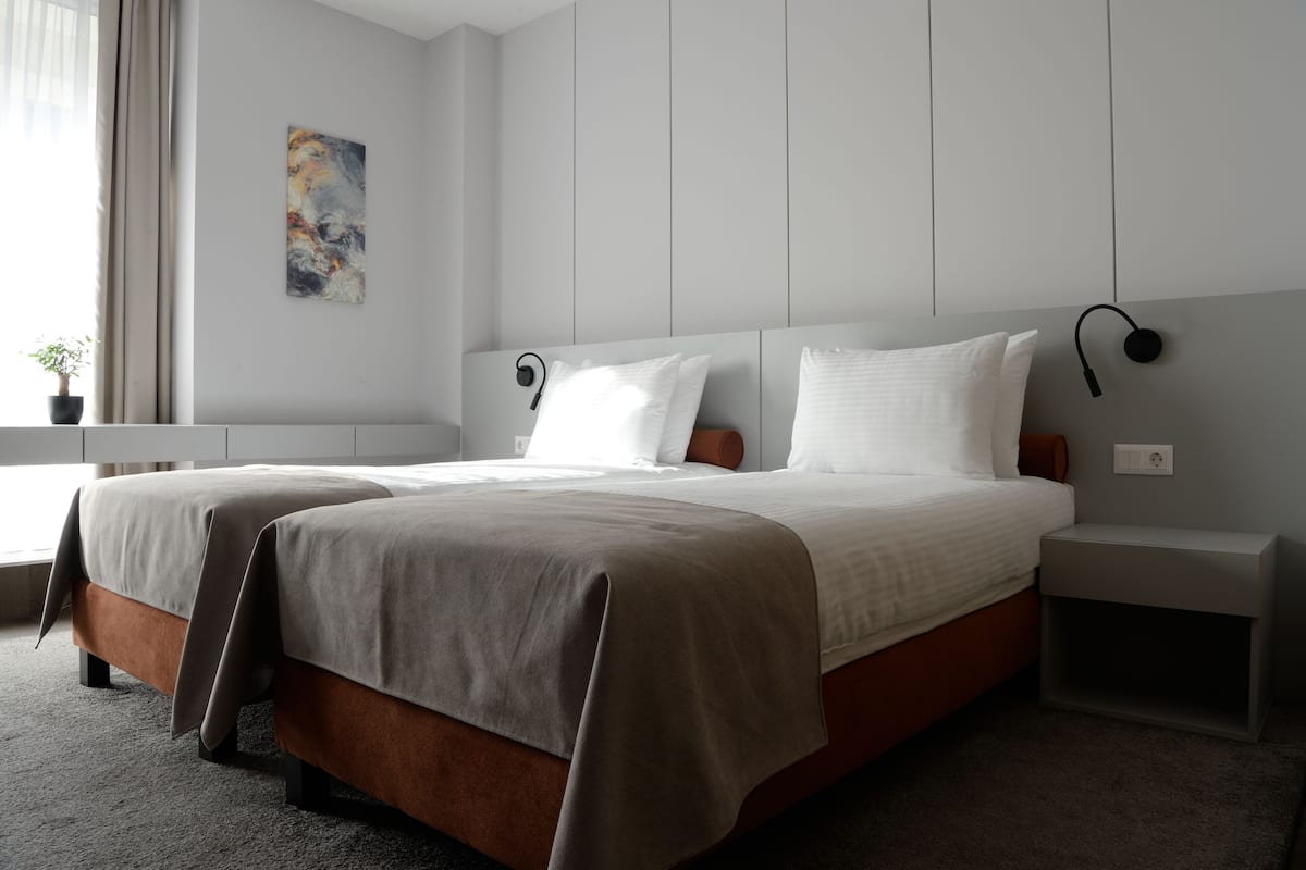 A modern hotel room with two beds, a bedside table, a nightstand, a wall-mounted lamp, and a window with curtains.