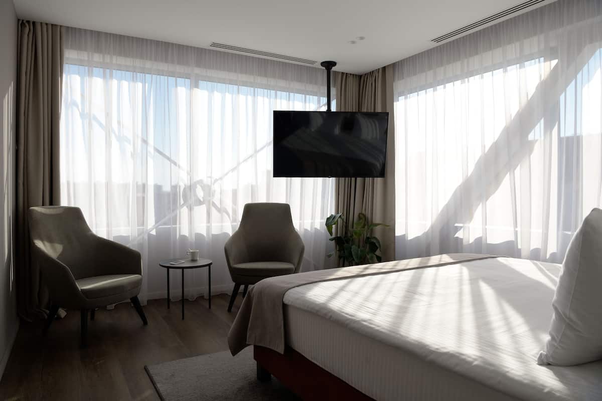 A hotel room with a bed, two armchairs, a small table, a TV, and large windows with sheer curtains.