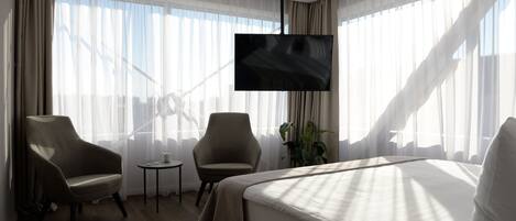 A hotel room with a bed, two armchairs, a small table, a TV, and large windows with sheer curtains.